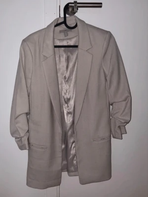 Beige oversized kavaj från H&M - Stilren beige oversized kavaj från H&M med klassisk krage och två fickor framtill. Kavajen har helfodrad insida i glansigt tyg och långa ärmar med lätt rynk vid ärmslut. 
