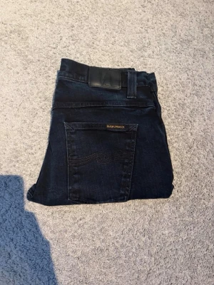 Nudie Jeans - Säljer ett par jeans från Nudie Jeans med klassisk femficksmodell och snygga detaljer på bakfickan. Jeansen har en rak passform och är tillverkade i mjukt denimtyg. Perfekta för dig som gillar stilrena och tidlösa jeans.midje mått 40cm längd 108cm