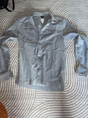 Ljusgrå skjorta/overshirt - Säljer en ljusgrå skjorta. Skjortan har två bröstfickor med knappar, klassisk krage och långa ärmar. Perfekt för dig som gillar en stilren och enkel look. Storlek XS/S och sprillans ny!