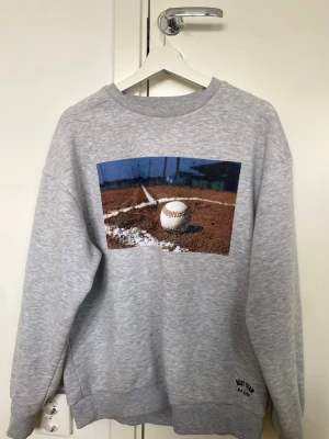 Grå sweatshirt med basebolltryck - Säljer en grå sweatshirt med ett stort basebollmotiv på framsidan och texten 'Remarkable Authentic EST 1998' i svart på ryggen. Tröjan har rund hals, ribbade muddar och är gjord i mjukt sweatshirtmaterial. Perfekt för dig som gillar sportig stil.