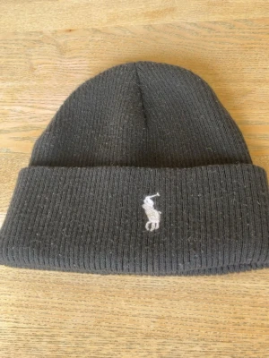 Svart ribbstickad mössa Polo Ralph Lauren - Säljer en svart ribbstickad mössa från Polo Ralph Lauren med klassisk uppvikt kant och den ikoniska broderade loggan framtill. Mössan är i mjukt material och har en enkel, stilren design som passar till det mesta.