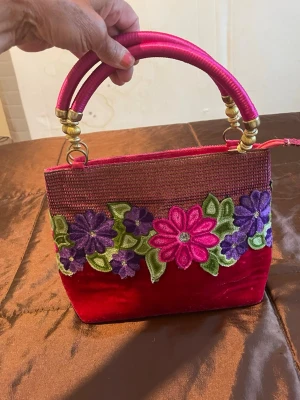 Rosa handväska med blommor - Unik handväska i starkt rosa tyg med broderade blommor i lila, rosa och grönt på framsidan. Väskan har två stadiga handtag med guldfärgade detaljer och en fyrkantig form. Materialet är en mix av tyg och sammet, vilket ger en lyxig känsla.handtaget är metall och nån sort band , 