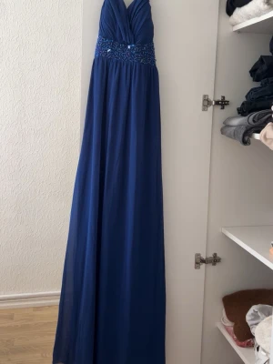Blå långklänning med broderad midja - Elegant blå långklänning med v-ringad omlottringning och broderade detaljer i midjan. Klänningen har en flowy passform och är ärmlös, perfekt för speciella tillfällen. Materialet känns lätt och följsamt.