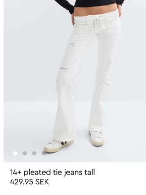 Vita bootcut jeans med snörning - Säljer ett par vita bootcut jeans med snörning i midjan och slitna detaljer på benen. Jeansen har hög midja och långa ben, perfekta för en trendig och avslappnad look. De är från gina