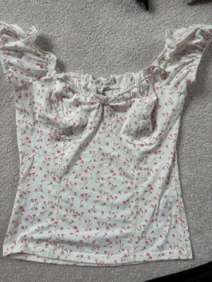Blommig topp med volang från NLY Trend - Supersöt topp från NLY Trend i vit med små rosa blommor. Toppen har volangdetaljer vid axlarna och en lätt rynkad byst. Perfekt för dig som gillar romantisk stil och vill ha något luftigt till sommaren. 🌸🌸