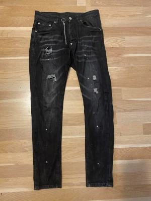 Svarta distressed jeans Dsquared2 - Säljer ett par svarta jeans från Dsquared2 med skinny fit, slitningar och hål på benen samt coola färgstänk. Jeansen har en unik dragkedja med ringdetalj och klassisk femficksmodell. Materialet är denim i bomull med stretch för extra komfort.