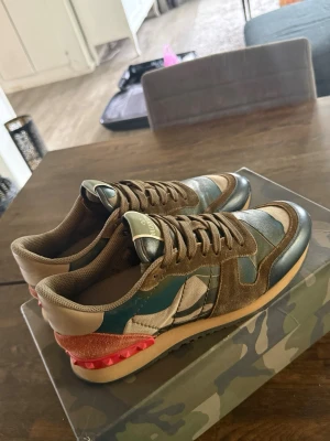 Valentino Garavani camo sneakers - Snygga sneakers från Valentino Garavani med camomönster i grönt, beige och brunt. Skorna har detaljer i mocka, läder och textil samt en röd hälflik med nitar. Klassisk låg modell med gummisula och bruna skosnören. Hör av er vid funderingar o kom med bud!😁Obs storlek 41 men passae även 42!