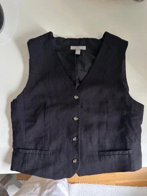 Svart väst från H&M med knappar - Stilren svart väst från H&M med fem knappar framtill och två fickdetaljer. Västen har en klassisk v-ringad skärning. 