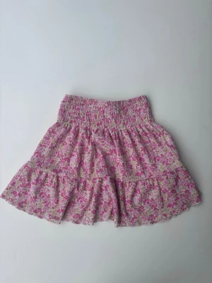 Blommig volangkjol från Gina Tricot - Supersöt kort kjol från Gina Tricot med blommigt mönster i rosa, vitt och grönt. Kjolen har bred resår i midjan och volangdetaljer som ger en luftig och trendig look. 