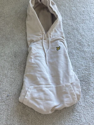 Beige hoodie från Lyle & Scott - Säljer en beige hoodie från Lyle & Scott med klassisk gul logga på bröstet. Hoodien har huva med dragsko, stor magficka och ribbade muddar vid ärmar och nederkant. Perfekt för en avslappnad och stilren look.