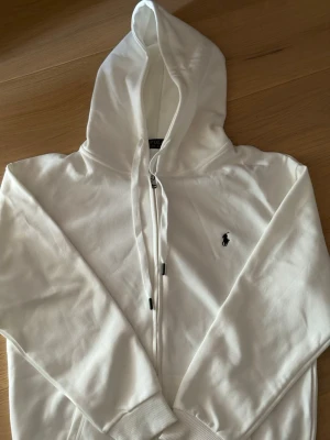 Vit hoodie från Polo Ralph Lauren - Snygg vit hoodie från Polo Ralph Lauren med dragkedja framtill och klassisk huva. Ikoniska lilla loggan broderad på bröstet. Tillverkad i mjuk bomull som känns skön mot huden. Perfekt för en clean och stilren look.