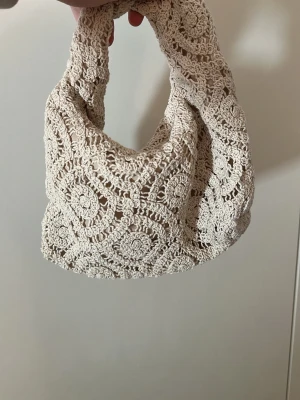 Clutch väska virkad  - Supersöt beige handväska i virkat material med rundade former och unikt mönster. Perfekt accessoar för dig som gillar boho-stil och vill ha något som sticker ut. Väskan har ett mjukt handtag och är lätt att bära.