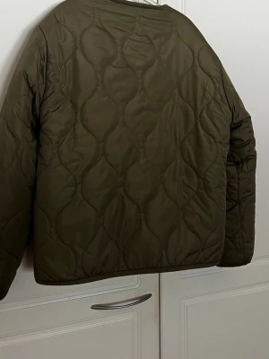 Olivgrön quiltad jacka - Säljer en olivgrön quiltad pufferjacka med knäppning framtill och två fickor. Jackan har en mysig beige fuskpäls på insidan och en oversized passform. Perfekt för kalla dagar och riktigt skön att bära. Passar dig som gillar en avslappnad och trendig stil.