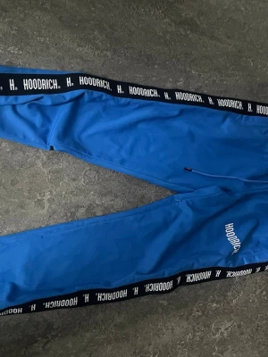 Blå mjukisbyxor från Hoodrich - Säljer ett par blå mjukisbyxor från Hoodrich med svarta sidor där det står HOODRICH i vitt. Byxorna har resår i midjan och raka ben. Perfekta för en chill och avslappnad stil. Materialet känns mjukt och bekvämt.