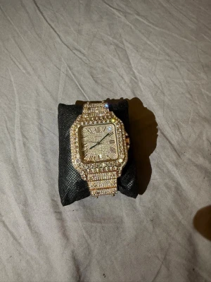 Ca**ier klocka iced out - Säljer en riktigt blingad klocka med fyrkantig urtavla och romerska siffror. Klockan är täckt av glittrande stenar och har ett armband i rosefärg. Perfekt för dig som vill sticka ut och älskar att glänsa.