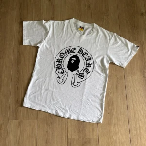 Vit Chrome Hearts x Bape T-shirt - Snygg vit t-shirt med stor svart Chrome Hearts x Bape-logga på bröstet. Klassisk passform och rund hals. Tillverkad i mjuk bomull som känns skön mot huden. Perfekt för dig som gillar streetwear och vill sticka ut med en unik design. 100% bomull