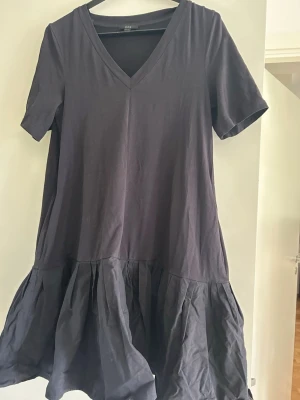 Svart kort klänning från COS - Svart kort klänning från COS med v-ringning och korta ärmar. Klänningen har en volang nedtill som ger en avslappnad och trendig look. Tillverkad i mjuk bomull, perfekt för dig som gillar enkel och stilren design. Märket COS