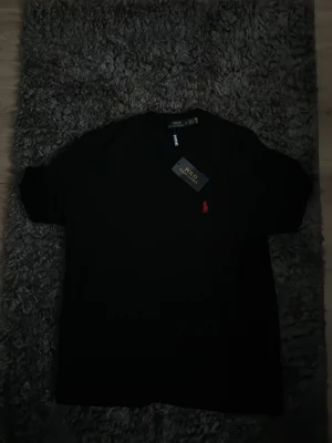 Svart t-shirt från Polo Ralph Lauren - Klassisk svart t-shirt från Polo Ralph Lauren med korta ärmar och den ikoniska röda logon broderad på bröstet. Tillverkad i mjuk bomull för en skön känsla och enkel stil. Perfekt till jeans eller shorts.