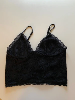 Svart spetsbralette med smala band - Trendig svart bralette i spets med tunna, justerbara axelband och v-ringad urringning. Spetsdetaljer längs kanterna och stretchig rygg för extra komfort. Perfekt för dig som gillar en feminin och modern stil.