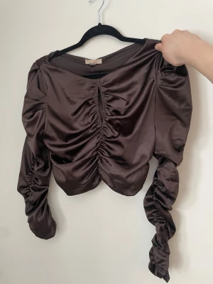 Brun satinblus med rynkade detaljer - Här har vi en av mina favoriter! svinsnygg, tight och lite urringad. Den är så pass tight ( i alla fall på mig) att man inte behöver ha bh på sig och man kan ändå dansa loss. Snygg drapering på armar, fram och bak. Har fått mycket komplimanger när jag haft på mig denna topp.  Armarna är lika långa, det är bara jag som inte dragit ut den ena armen ordentligt. Snygg brun färg.