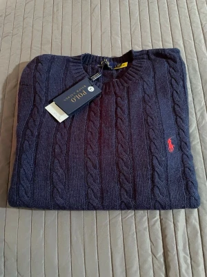 Mörkblå kabelstickad tröja Polo Ralph Lauren - Kabelstickad tröja i mörkblått från Polo Ralph Lauren med rund halsringning och röd broderad logga på bröstet. Tröjan har ribbade muddar vid ärmslut och nederkant och är tillverkad i mjukt material. Perfekt för en stilren och klassisk look.