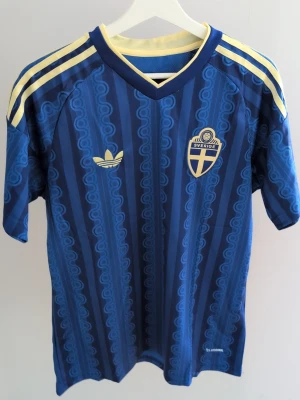 Blå Sverige matchtröja Adidas - Finns 3 stycken Helt ny  matchtröja från Adidas med Sveriges landslagsemblem på bröstet och gula detaljer. Tröjan har korta ärmar, V-ringning och klassiska Adidas-ränder på axlarna. Tillverkad i lätt och ventilerande syntetmaterial, perfekt för fotboll eller samling.