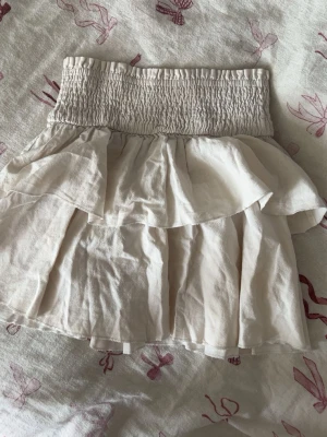 Beige volangkjol med smock - Söt beige kortkjol med bred smock i midjan och dubbla volanger. Kjolen har en luftig och lätt känsla, perfekt för varma dagar. Passar till både sneakers och sandaler för en avslappnad look.