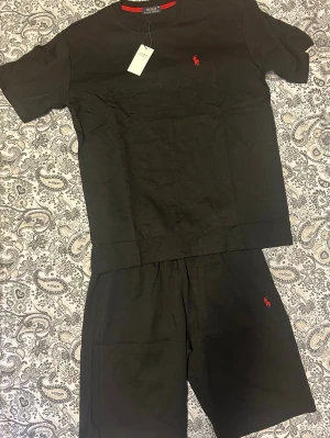 Svart set från Polo Ralph Lauren - Säljer ett svart set från Polo Ralph Lauren med t-shirt och shorts. Båda plaggen har den klassiska röda loggan broderad på bröstet och benet. Setet är gjort i mjukt bomullsmaterial och har en avslappnad passform. Perfekt för chill dagar eller som snyggt loungeplagg.