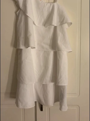 Vit volangklänning från Gina Tricot - Säljer en vit klänning från Gina Tricot med volangdetaljer och asymmetrisk axel. Klänningen har en luftig passform och är perfekt för sommarens alla tillfällen. Tillverkad i ett lätt material som känns skönt mot huden.