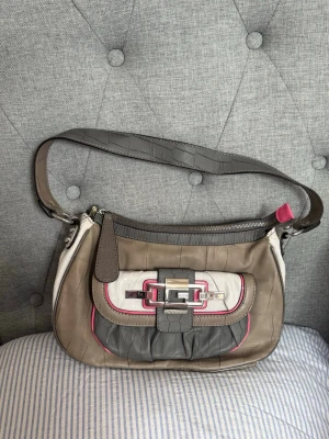Guess axelremsväska i beige och grå - Äkta handväska från Guess i beige, grå och vitt med detaljer i rosa. Väskan har ett coolt spänne framtill och präglat mönster på materialet. Axelremmen är i matchande grått och insidan har Guess-logga överallt. Har lite slitage på sidan av remmen (syns på fotona)