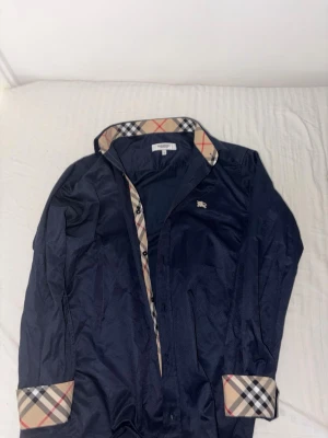 Mörkblå Burberry skjorta med rutor - Snygg mörkblå skjorta från Burberry med klassiskt rutigt mönster på insidan av krage och manschetter. Skjortan har långa ärmar, knappar framtill och en liten broderad logga på bröstet. Perfekt för dig som gillar stilrena detaljer och ikonisk design.