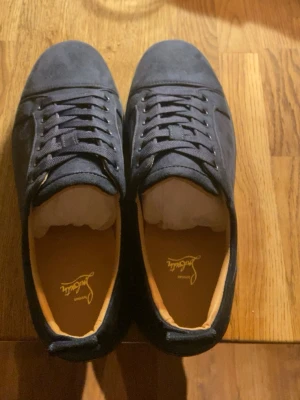 Mörkblå sneakers från Louboutin - Snygga mörkblå sneakers från Christian Louboutin i mocka med klassisk röd sula och diskreta detaljer. Lågt skaft och matchande blå skosnören. Insidan är ljusbrun med guldfärgad logga. Perfekta för dig som gillar exklusiv streetstyle. Går att pruta ner litegrann. 