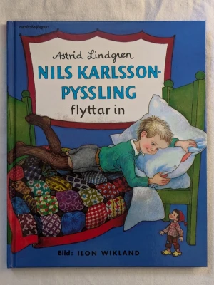 Nils Karlsson-Pyssling flyttar in - En färgglad bilderbok av Astrid Lindgren med illustrationer av Ilon Wikland. Omslaget visar en pojke på en säng med ett lapptäcke i olika färger och en liten figur på golvet. Boken har hårda pärmar och tydliga illustrationer.