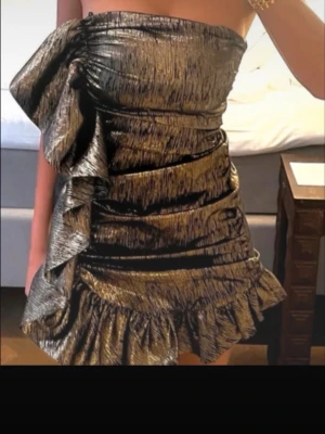 Super trendig klänning ifrån Zara!! - Säljer denna super populära och trendiga klänning från Zara som är helt slutsåld. Klänningen har ett band men går även att använda axelbandslös som på bild 1!🥰Snygg kort klänning i guldig metallic med volangdetaljer och asymmetrisk design. Klänningen har en unik struktur och passar perfekt för dig som vill sticka ut.