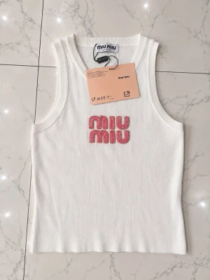 Vit linnetopp från Miu Miu - Snygg vit linnetopp från Miu Miu med rosa pärlbroderad logotyp på bröstet. Toppen är ärmlös och har en ribbad struktur som ger en modern och clean look. Perfekt för att skapa en trendig outfit.