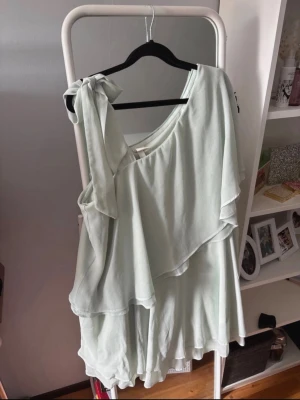 Ljusgrön oneshoulder klänning H&M - Säljer en ljusgrön oneshoulder klänning från H&M med volangdetaljer och knyt på axeln. Klänningen har en luftig och flowy passform, perfekt för varma dagar. Tillverkad i ett mjukt och lätt material som känns skönt mot huden.