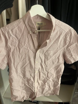 Rödvit randig skjorta East West - Kortärmad skjorta från East West med smala röda och vita ränder. Klassisk button-down krage och knäppning framtill. Tillverkad i bomull, perfekt för varma dagar. Enkel bröstficka och avslappnad passform.