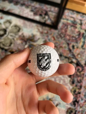 Golfboll Delsjö Golfklubb - Vit golfboll med Delsjö Golfklubbs logga i svart och vitt tryck på ena sidan och texten 'TILLHÖR' på andra sidan. Klassisk rund form med dimple-mönster för bättre flygförmåga. Perfekt för dig som gillar golf och vill ha en boll med klubbkänsla.