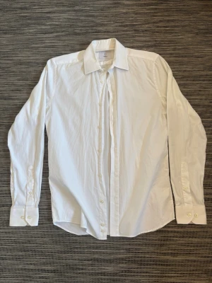 Vit klassisk skjorta från Bläck - Säljer en stilren vit skjorta från Bläck med klassisk krage och långa ärmar. Skjortan har knappar framtill och vid ärmsluten, perfekt för en clean och tidlös look. Tillverkad i mjuk bomull som känns skön mot huden.