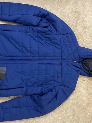 Blå dunjacka från Peak Performance - Snygg blå dunjacka från Peak Performance med huva och dragkedja framtill. Jackan har en bröstficka med dragkedja och diskret logga på bröstet. Perfekt för kalla vinterdagar tack vare det värmande dunmaterialet och den sportiga designen.