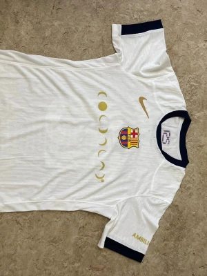 Vit FC Barcelona matchtröja Nike - Säljer en vit FC Barcelona matchtröja från Nike med guldfärgade detaljer, klubbmärke på bröstet och månfas-tryck. Svarta muddar vid ärmar och hals. På ryggen står det "Lamine Yamal" och nummer 10 i gult samt UNHCR-logga. Perfekt för dig som älskar fotboll och Barca.