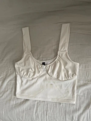 Vit croppad linnetopp från H&M - Säljer en vit croppad linnetopp från H&M med breda axelband och lätt rynkade detaljer vid bysten. Toppen är ärmlös och har en enkel, stilren design som passar perfekt till sommaren. Materialet känns mjukt och stretchigt.