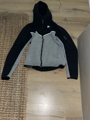 Nike hoodie svart och grå XS - Snygg hoodie från Nike i svart och grått med dragkedja framtill och vit detalj vid bröstet. Tröjan har huva, långa ärmar och en ficka med dragkedja på ena ärmen. Perfekt för en sportig och avslappnad stil.