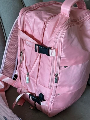 Stor rosa ryggsäck med många fack - Rymlig rosa ryggsäck med flera dragkedjeförsedda fack och justerbara axelband. Väskan har svarta spännen och mesh på baksidan för extra komfort. Perfekt för dig som vill ha plats för allt och gillar en färgglad stil.