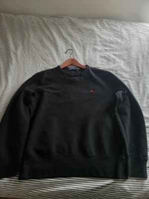 Svart sweatshirt från Polo Ralph Lauren - Klassisk svart sweatshirt från Polo Ralph Lauren med rund halsringning och diskret röd logobrodyr på bröstet. Tröjan har ribbade muddar vid ärmslut och nederkant. Perfekt för en avslappnad och stilren look.