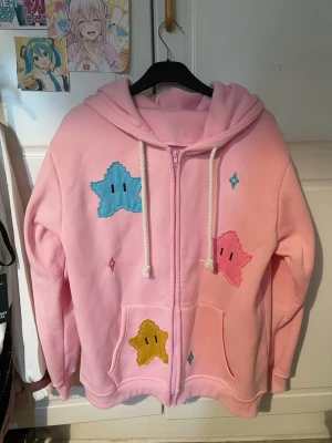 Rosa hoodie med Super Mario stjärnpatchar - Säljer en ljusrosa hoodie med dragkedja och huva. Hoodien har stora broderade stjärnpatchar i blått, gult och rosa på framsidan samt vita snören i huvan. Mjuk och skön bomullsmix. Köpt från Temu (jag vet 😥men det var 2023 och jag var fattig) Original pris ca 250~300kr. Säljer för 180kr. Storlek L-XL (oversized) Knappt använd