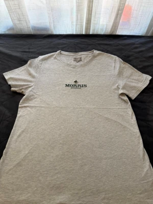 Ljusgrå t-shirt från Morris - Säljer en stilren ljusgrå t-shirt från Morris med diskret logga och text i grönt på bröstet. Klassisk passform med rund hals och korta ärmar. Perfekt basplagg i mjuk bomull som funkar till det mesta.
