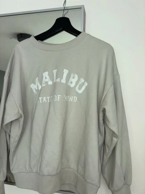 Ljusgrön sweatshirt med MALIBU-tryck - Säljer en ljusgrön sweatshirt med texten 'MALIBU STATE OF MIND' i vitt framtill. Tröjan har rund halsringning, ribbade muddar vid ärmslut och nederkant samt en avslappnad passform. Perfekt för en chill och casual stil.