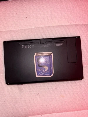 Nintendo Switch svart konsol - Säljer en svart Nintendo Switch konsol med en stor skärm och klassisk rektangulär form. Baksidan har en unik klistermärke med motiv. Konsolen är tillverkad i plast och har tydliga Nintendo-loggor. Perfekt för gaming hemma eller på språng.