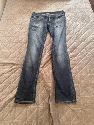 Blå bootcut jeans från Nudie Jeans - Säljer ett par blå bootcut jeans från Nudie Jeans med slitningar på låren och vid fickorna. Jeansen har klassisk femficksmodell, orangea sömmar och knappgylf. Perfekta för dig som gillar en avslappnad och trendig stil.